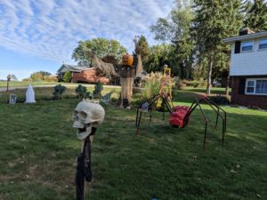 Our front yard decorated for Halloween. A skull tiki torch in the foreground, a giant red spider, a scarecrow, and the creepy white lady. A blue sky with a few clouds overhead.