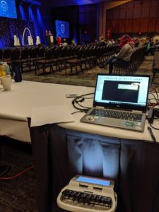 A steno machine under a table, a laptop on the table, and a view of the stage at Wordcamp US.