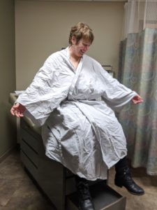 Amanda laughing while wearing a gown that goes all the way to her ankles, sitting on the doctor's table, holding her arms out to show how giant the robe is.