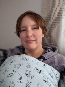 Selfie: At home wearing a soft purple jacket with black on it. My mastectomy pillow is in the foreground - it's blue with little dog pictures all over it.