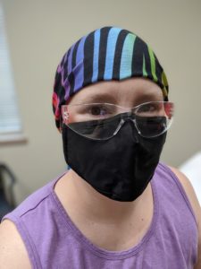 Selfie: black mask, safety glasses, purple tank top, zebra-striped head covering with rainbow colors and black stripes.