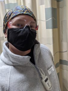 Selfie: black mask, safety glasses, gray jacket with Purell hanging from the zipper, a sticker that says: screened and the date. A head covering with muted colors and abstract design.