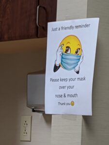 A sign that says: Just a friendly reminder - please keep your mask over your nose and mouth. It has a picture of a smiley face with arms wearing a mask.