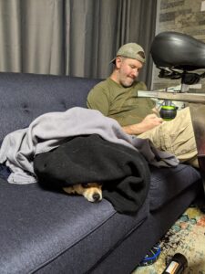 Arthur on our blue couch under a bunch of blankets. Only his nose is poking out a bit. Gabe, my husband, is sitting on the couch next to him. You can also see the back of our spinning bike that we keep in that room randomly. 