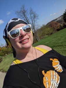 Selfie out for a walk wearing a Halloween shirt that is two skeleton holding pumpkins over where my tatas used to be. I'm also wearing a skull buff on my head and sunglasses.