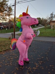 Gabe dressed as a pink unicorn in an inflatable suit. Rainbow mane and tail. Holding an axe and a beer.