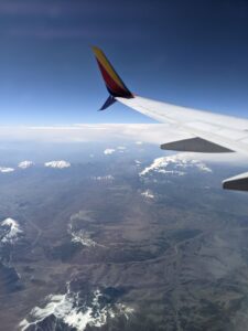 Looking out the airplane window. Part of the wing is visible. Snowcapped mountains in the distance and a few low clouds.