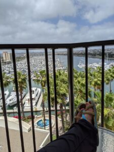 I'm relaxed on my balcony, you can see my feet up on the table. I'm wearing rope sandals that are brown and blue. The rail of the balcony is in front of me and beyond that is the marina with all the boats moored. There are palm trees closer in.