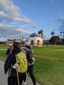 Blue sky with some clouds, a park with green grass with a domed open-air building. The dome is blue tiles with other colors, hard to see from this angle. There are a few other Athenas walking ahead of me.