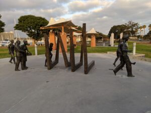 An outside statue installation. There are bronze squares that form a tunnel and people are behind, one person is entering, and a family is on the other side, having made it through the trials of cancer. All in bronze.