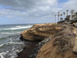 A shoreline of the Pacific Ocean. The cliffs are brown rock/sand and rise above the water. There are palm trees on top. The sky is clouds with some blue peeking through. The ocean is churning against the rocks.