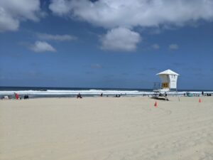 A beach on the Pacific Ocean. Rolling waves coming in, blue sky with a few clouds. There's a lifeguard booth on stilts near the right side of the photo.