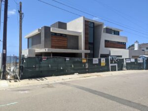 A giant, squarish modern house. It's light brown with darker brown aroudn the window areas, lots of windows. It's surrounded by a construction fence.