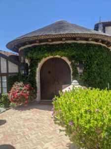 A house entrance. It is round with a curved doorway, like you'd find in a castle, but much bigger. There is ivy growing all over it. The sky is blue, there are some native plants in the foreground.