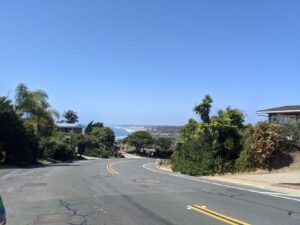 Walking down from Mt. Soledad on a road. There are trees and bushes and sand along the side of the road. The Pacific Ocean is in the distance. The sky is very blue.