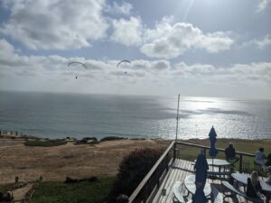 Evening. The sky is clouding up again. The Pacific Ocean is reflecting the light of the sun. A few gliders are in the air and we are looking out off of a balcony overlooking it all.