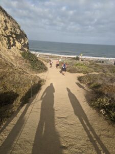 Walking down a sandy trail, a hill to the left and the Pacific Ocean in front of us. Clouds with blue sky peeking out. You can see our shadows - three of us, - stretching out in front of us and a few Athenas further down the trail.