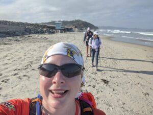 Selfie. I'm smiling a little, wearing sunglasses and my Athena buff. Lucy and the reaper trail behind me. The Pacific Ocean is on the right and we are walking on sand. The sky is cloudy.