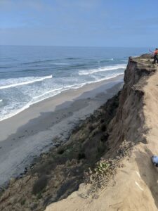 A cliff down to the Pacific Ocean. It's probably all made of sand, so... be careful. The waves are hitting the shoreline. An Athena is ahead taking a picture out over the ocean. Far ahead. Blue sky with a few clouds.