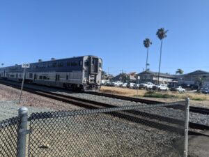 A train on the tracks to the left heading to the right, moving fast, at a controlled intersection. Blue sky. It's a commuter train I think.