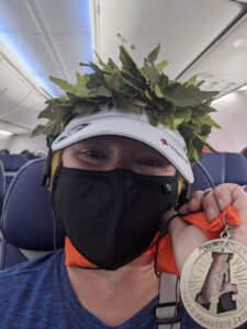 Selfie on a plane. I'm wearing my Athena Adventures hat, but over it is the wreath we got at the end. I'm holding up my medal. I'm wearing a black mask. I'm smiling though.