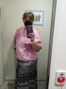 A picture of me wearing a pink wrap around top that is used for breast exams and a black and white patterned long skirt. My hair is short and I'm wearing a black mask.