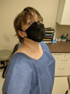 Selfie wearing a blue paper top that opens in the back. I'm wearing a black mask and looking up at the camera, hair in my eyes. It's short, but I do need a haircut.