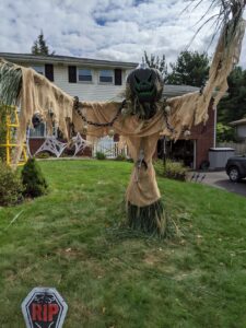 A scarecrow made of burlap and long grass. It has skulls hanging from thick chains on its arms, a skeleton on its front. The head is spray painted black with green on the eyes, nose, and mouth and looks like it's dripping. The stem is spray painted silver.