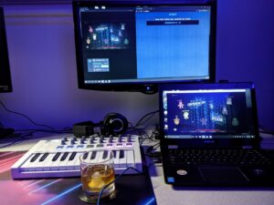 A little keyboard and a glass of whiskey. There is a laptop and a screen above it. The lighting is purple-ish.