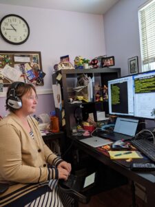 Amanda hard at work. I've got a bunch of screens, a laptop, a steno machine. I'm dressed in a 'work outfit' of a striped dress and a brown blazer to stay warm. There's a clock on the wall and lots of random weird stuff on my bulletin board