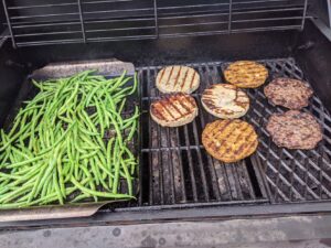 A grill with fresh green beans on the left on a pan and meat patties - chicken and hamburger - on the right.