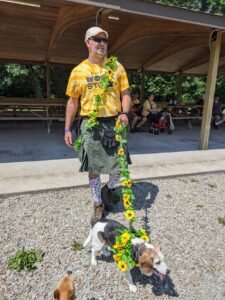 Gabe dressed up for Woofstock in a kilt, tall dog print socks, a yellow Woofstock shirt, and a flat cap. Edie is also dressed up with sunflowers all over her harness and up her leash. They're stepping out to lead the parade.