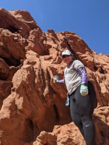 Amanda standing by some rocks dressed in black leggings, a gray shirt, purple arm sleeves with some white and black on them, green fingerless gloves with black flowers on them, sunglasses and Athena hat in white. It was hot. I kinda look like I'm holding up the rock.