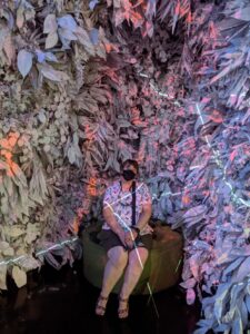 Amanda seated on a bench at a Meow Wolf art installation. It's kind of like a weird cave with lights all over. Amanda is wearing a green skort, a white shirt with purple flowers and sandals. And a black mask because inside/COVID!