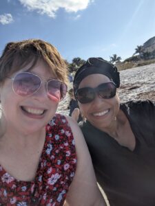 Amanda and Lucy taking a selfie. Amanda in the red dress with flowers and Lucy in black. We are being photobombed a bit by her little girl. We are sitting on Indian Rocks Beach in Tampa and the sky is blue with some white fluffy clouds.