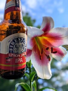 A bottle of Firestone Union jack IPA by an Asiatic lily that's white petals with pink on the inside. Blue sky in the background. 