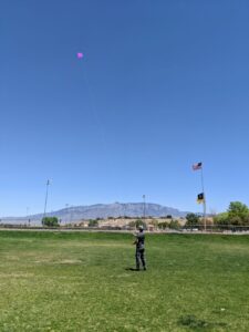 Gabe flying a purple kite in a huge open green space with the Sandia Mountains in the distance. Blue sky.