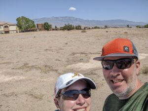 Selfie of Amanda and Gabe in the desert, the Sandia Mountains in the background with one little cloud in the blue sky.