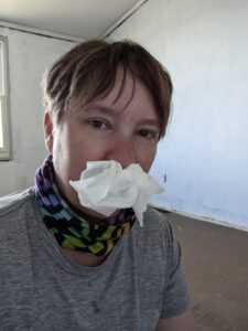 Amanda selfie with Kleenex shoved up my nose. I have a rainbow and black buff around my neck and my hair is short and kind of limp. I look a little rough. This is construction while having COVID. Yay. Gray shirt. Matched my mood.