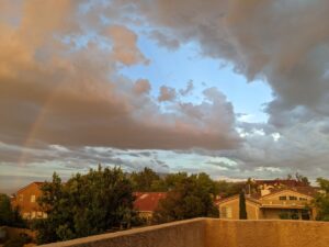 From our upstairs balcony, a shot of the sky with half a rainbow to the left, clouds starting to separate above, and more clouds to the left. The neighborhood is green and the Sandia Mountains are in the distance.