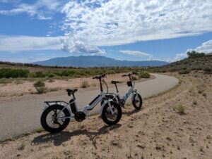 Two Lektric ebikes, both white with blue lettering and fat tires. They're on the side of a bike path with the Sandia Mountains in the distance. The sky is blue with quite a bit of clouds, but they're arrayed like a fan. Another gorgeous day.