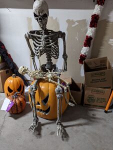 A skeleton sitting on a giant pumpkin head in the garage. The skeleton is holding a little skeleton on its lap. There are more pumpkins on the floor as well.