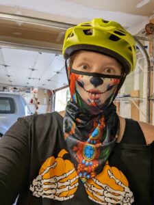 Selfie of me wearing a shirt that has skeleton hands holding pumpkins where my breasts used to be. I'm wearing a sugar skull face covering and have on a bike helmet. Yes, I dressed up to ride bikes!