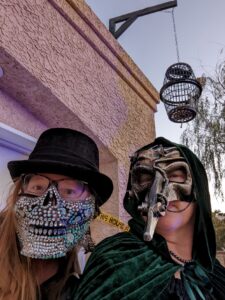 Selfie of me and my sister. She's wearing a big glittery half skull mask and a top hat. I'm wearing a steampunk palgue doctor mask and a cloak.  Behind us is the other gallows hanging over the garage. It just is full of bones in the bottom and sticking out.