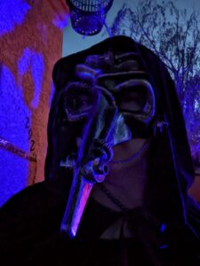 Selfie wearing my plague doctor mask with blacklights behind me. Head covered by a cloak.