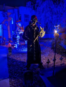 Gabe dressed in his half mask, a hat, a vinyl apron, holding an axe, standing in front of the Halloween decor with a blacklight at night.