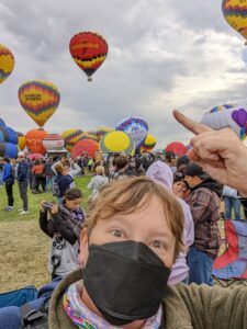 Selfie of me at the Balloon Fiesta. I'm wearing a mask, my hair is a mess, and I'm pointing up and over my head to a balloon that says 'Visit Albuquerque' - and there are other balloons inflating near the ground as well.