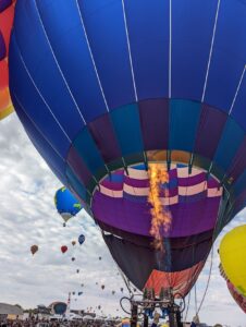 A blue hot air balloon being filled with hot air. The flame is on going up into the balloon. There are more going up in the background.