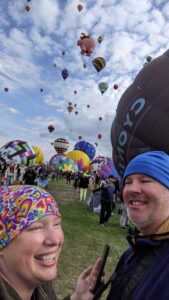 Amanda and Gabe at the Balloon Fiesta. Amanda is laughing and wearing a multi-colored head covering, Gabe is smiling and wearing a blue beanie. There are hot air balloons going up in the air behind us and all around us.