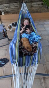 Amanda in a hammock in the backyard with blankets and Arthur on top. He loves hammock time.